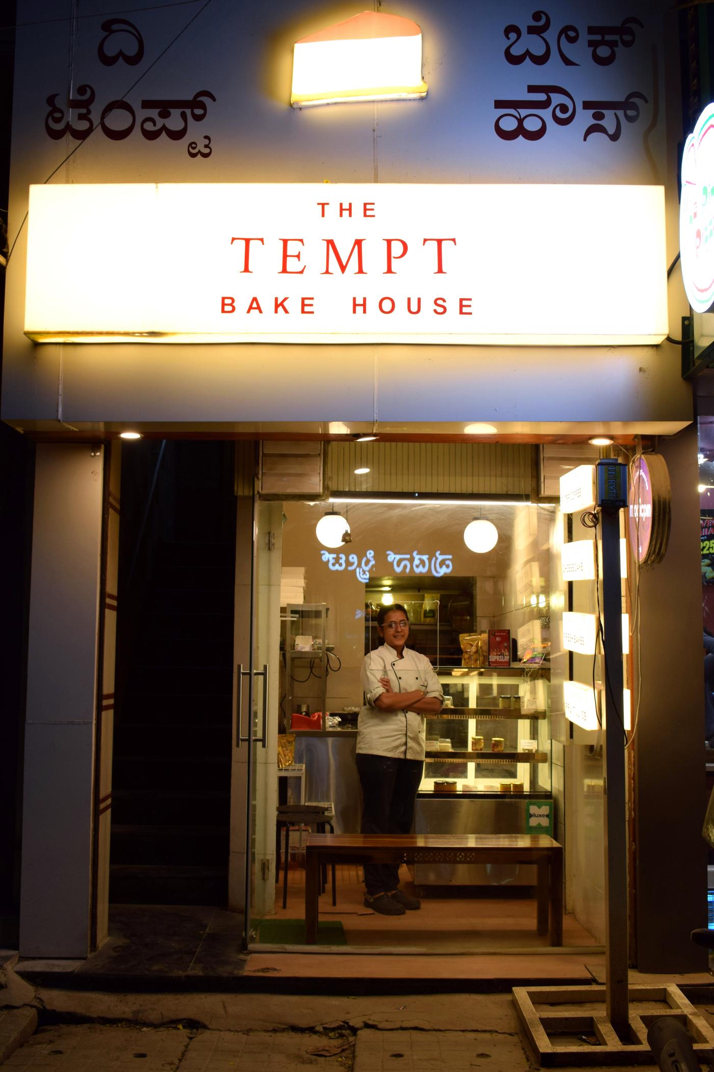 The Tempt Bake House at night, before it closed: the lit red sign, Kannada lettering above it, and Sneha standing inside the tiny glass-fronted bakehouse on 80 Ft Road, Koramangala
